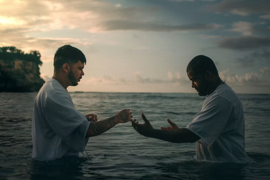 Two men waist-deep in water facing each other, one with hands extended as to performing a baptism ceremony