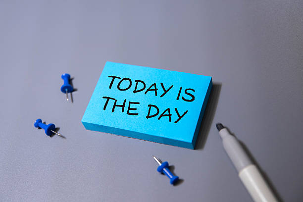 Blue sticky note with text 'TODAY IS THE DAY', surrounded by blue push pins and a marker on a gray surface