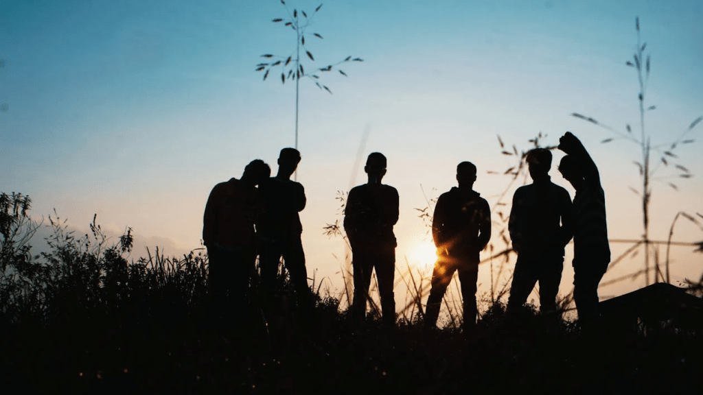 Silhouette of a group of young people standing in a field during sunset, with a colorful sky in the background.