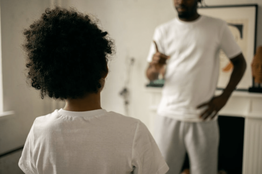 A father and child interact at home, with the father gesturing while giving instructions to the child, who has curly hair and is facing away.