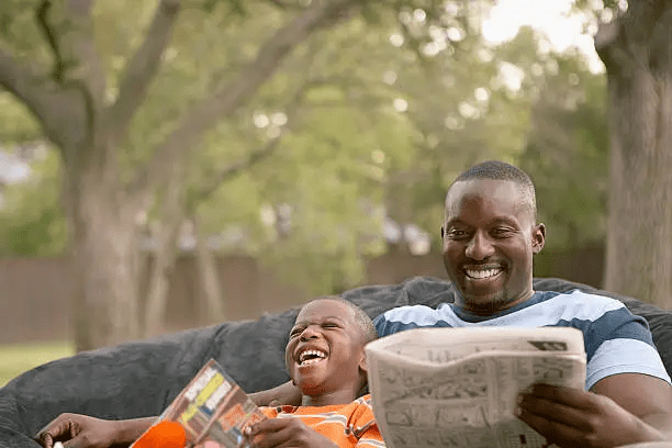 A father and son sitting together on a couch outdoors, both smiling and laughing while reading magazines.