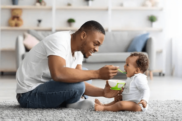 A father feeding his baby on a rug in a bright and cozy living room.