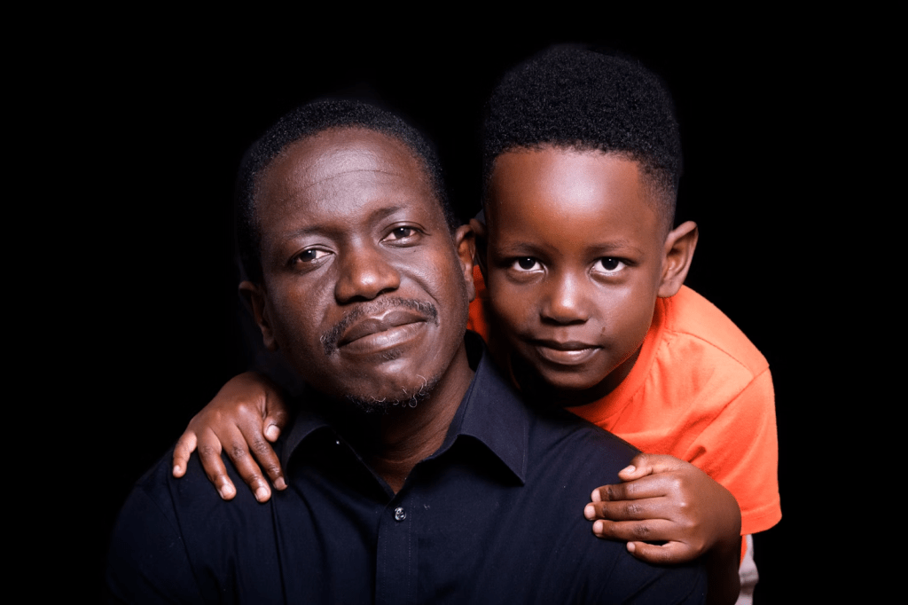 A father and son pose closely together, with the son hugging his father from behind. The father has a gentle smile, and both are against a dark background.