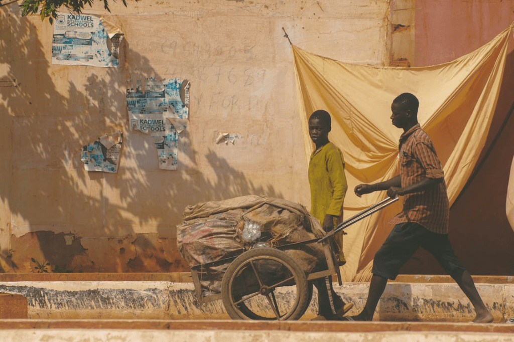 A father and a son walking alongside a cart filled with bags, with a textured wall and fabric background.