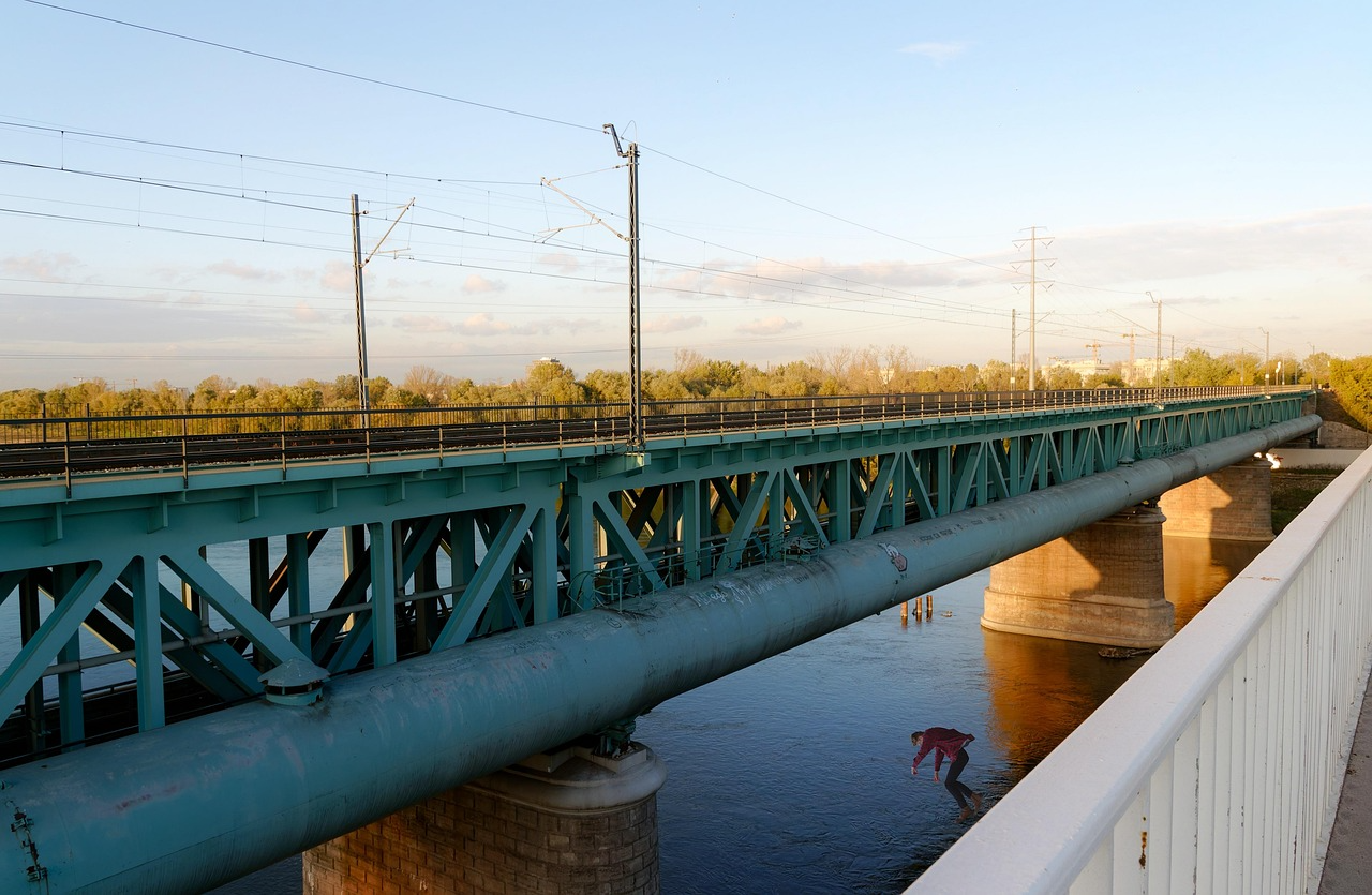 Turquoise railway bridge with overhead wires, pipeline, river, and person below
