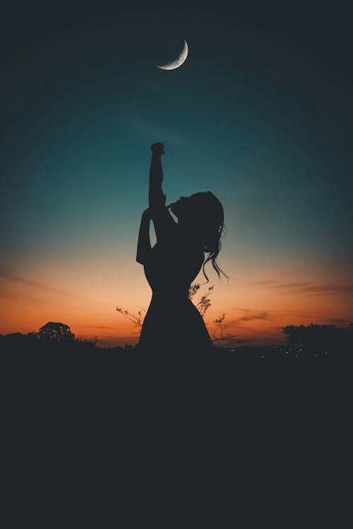 Silhouette of a woman reaching towards a crescent moon against a vibrant sunset sky.