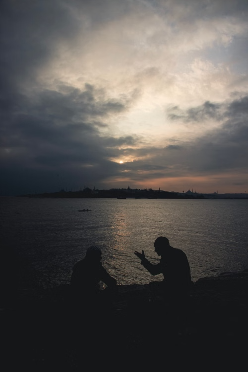Silhouetted conversation at sunset by water Two people sitting by the water at sunset, one gesturing with hands