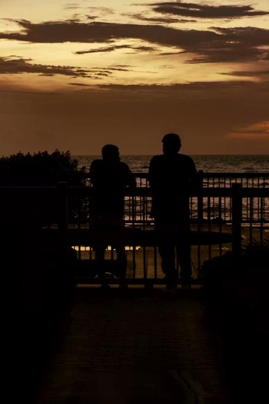 Sunset silhouette of two people by railing Two people standing by a railing looking at the ocean during sunset with clouds in the sky