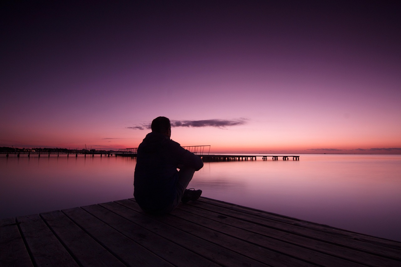 Contemplative silhouette at dusk pier Person sitting on a wooden dock watching a calm lake at sunset with a purple and orange sky