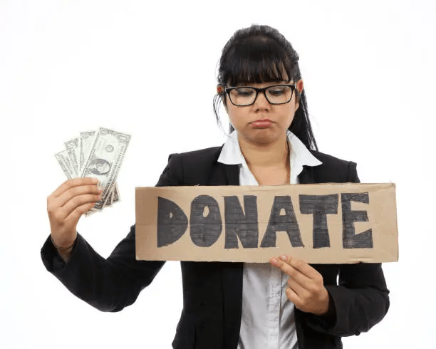A woman in a black blazer holds a sign that reads 'DONATE' while looking disheartened, with several dollar bills in her other hand.