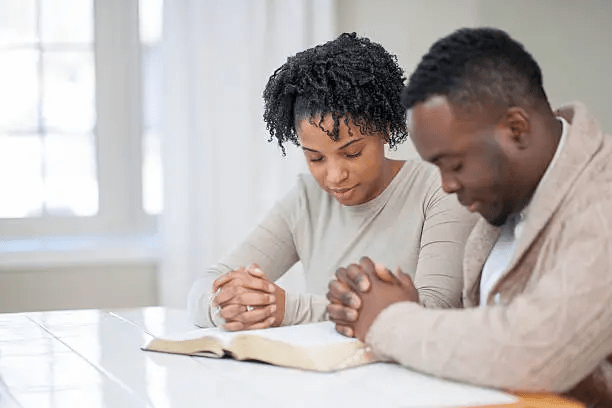 A man and a woman are sitting at a table with a Bible open in front of them, engaged in prayer with their heads bowed and hands clasped.