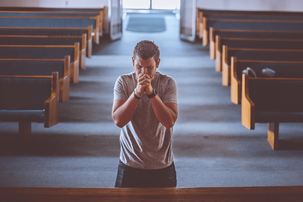 A man praying in a church setting, with wooden pews visible in the background.