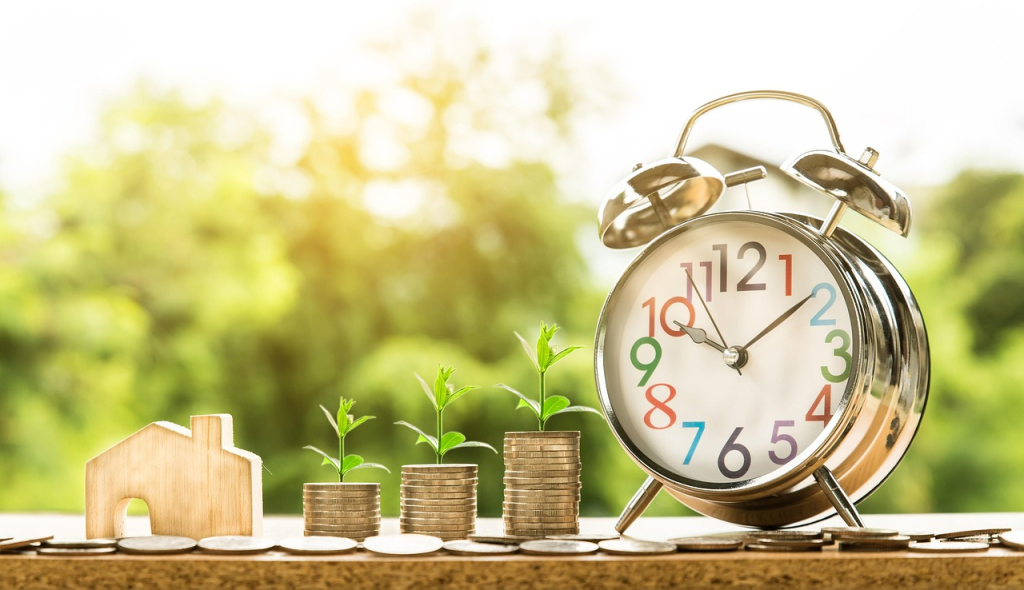 A wooden house symbolizing home ownership, alongside stacks of coins and small green plants, with a silver alarm clock in the background, representing financial growth and time management.