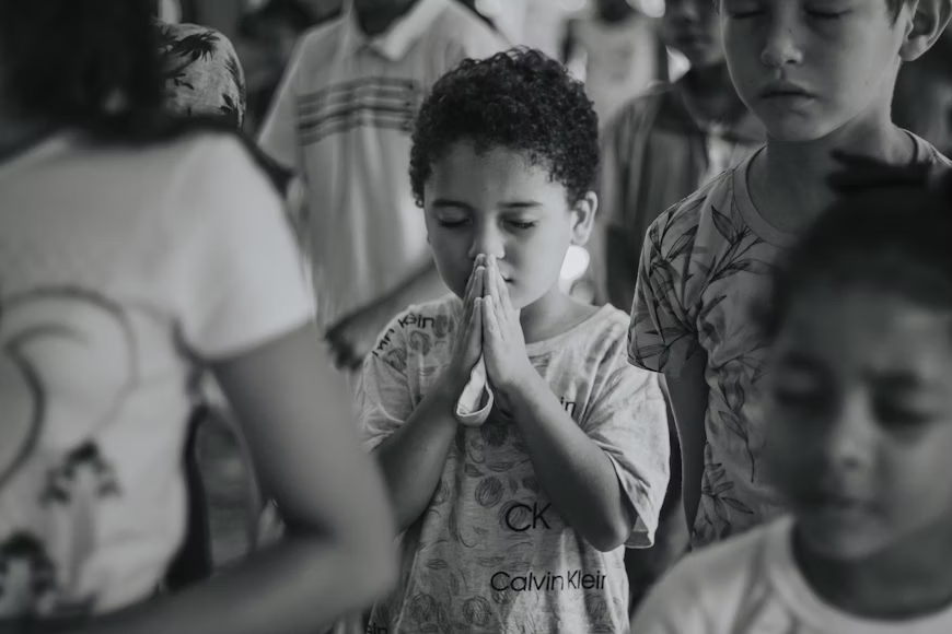 A young child with curly hair is shown with hands clasped in prayer, surrounded by other children who have their eyes closed. The image is in black and white, emphasizing the child's serious expression during a moment of reflection.
