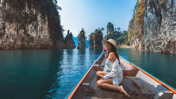 A woman sitting on a boat, wearing a white shirt and hat, enjoying a scenic view of tall rock formations and calm water in a beautiful landscape.