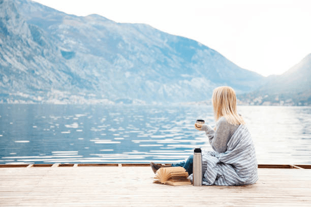 A person sitting on a wooden dock, wrapped in a blanket, enjoying a hot beverage while reading a book by a serene lake surrounded by mountains.