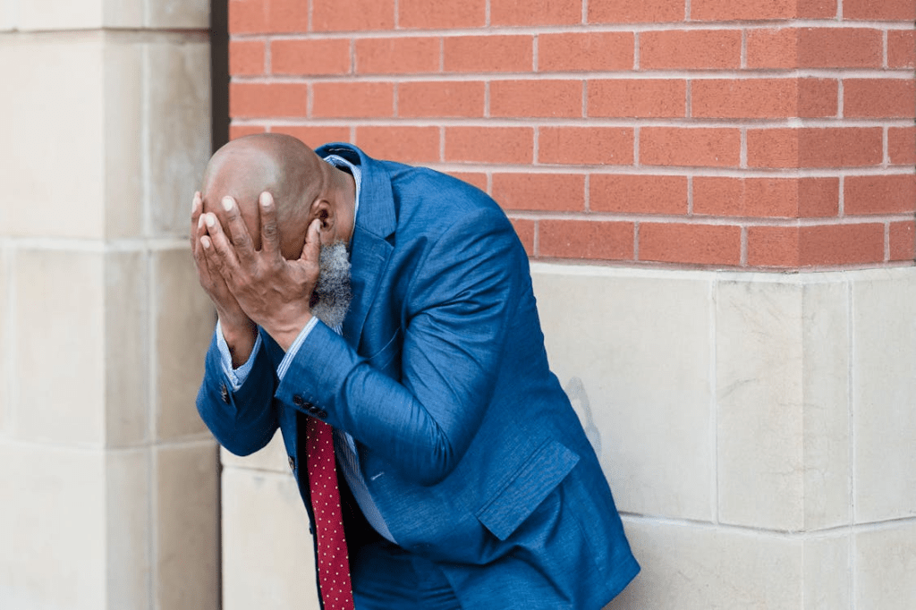 A man in a blue suit is leaning against a wall, covering his face with his hands, conveying a sense of distress or frustration.