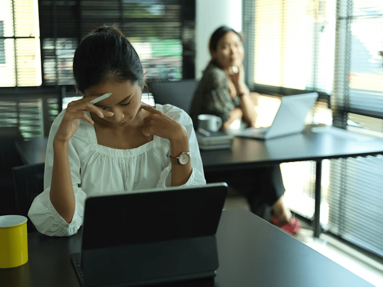 A woman in a white blouse is seated at a desk, appearing pensive while looking at her laptop. Another woman can be seen in the background, glancing towards her.