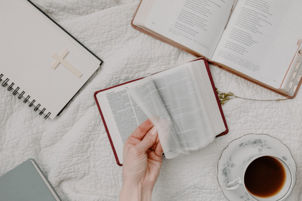 A hand flipping through the pages of an open Bible, accompanied by a notebook with a cross symbol, a cup of tea, and a potted plant, all placed on a textured white surface.