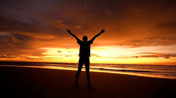 Silhouette of a person standing on a beach at sunset, with arms raised towards the sky.