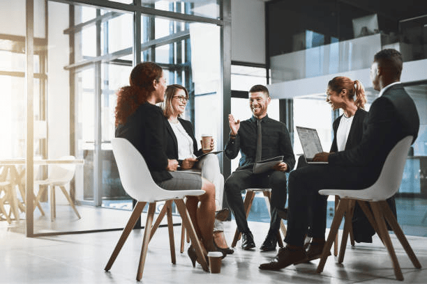 A group of five professionals engaged in a discussion in a modern office setting, with large windows allowing natural light to fill the room.