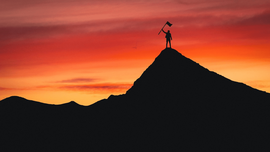 Silhouette of a person standing triumphantly on a mountain peak at sunset, holding a flag.