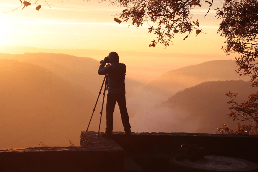 A silhouetted photographer standing on a rocky ledge, capturing the sunrise over misty hills.