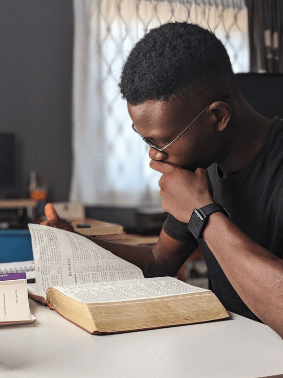 A person reading a Bible intently while sitting at a table, with natural light coming from a window.