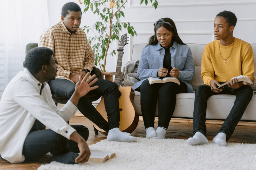 A group of four people engaged in a discussion, sitting on a rug in a cozy living room, with two individuals holding open Bibles.