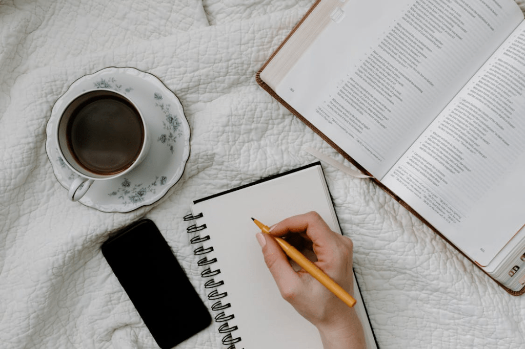 A hand writing in a notebook beside an open Bible, a cup of coffee, and a smartphone on a white blanket.