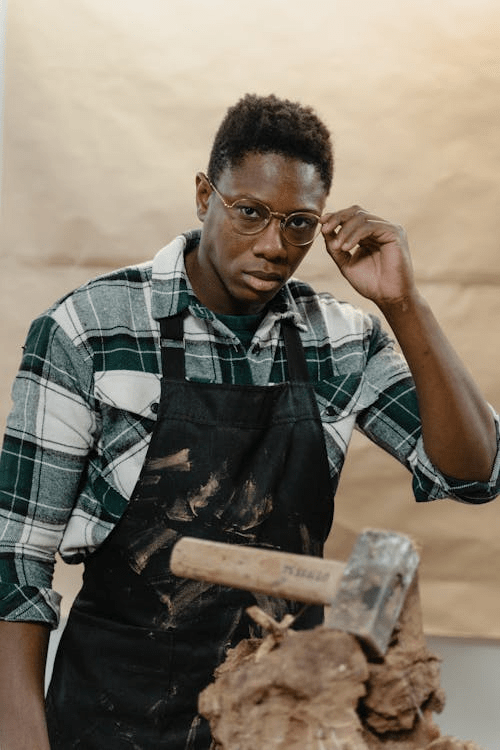 A focused artist wearing glasses and a flannel shirt works on a clay sculpture, holding a tool in one hand, amidst a workspace with clay materials.