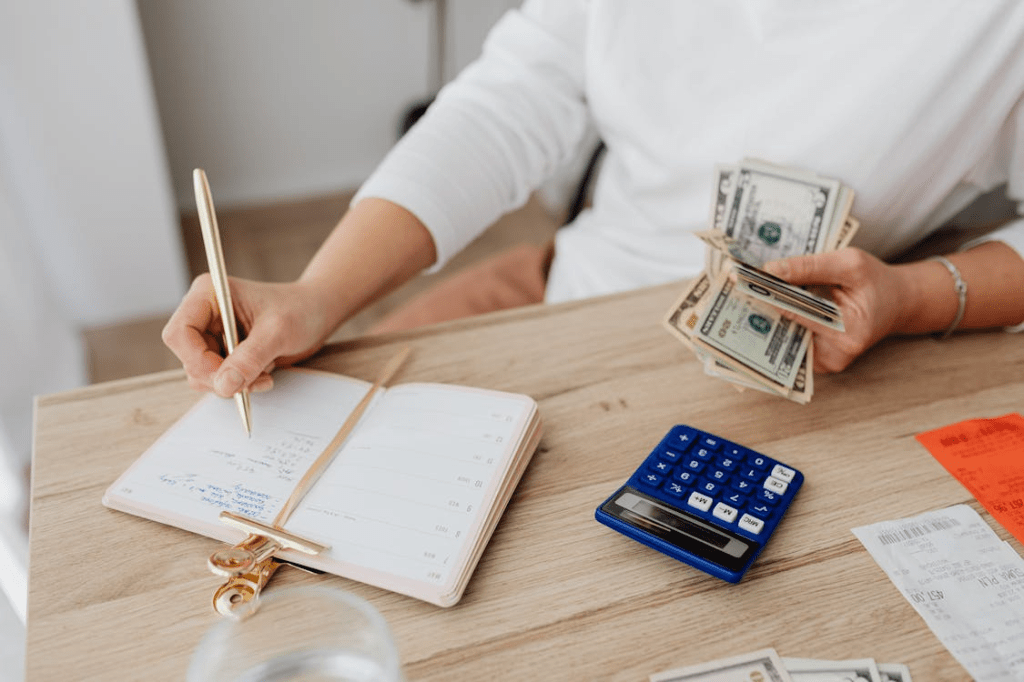 A person sitting at a wooden table, writing in a planner with a gold pen while holding cash in one hand. A blue calculator and receipts are also visible on the table.