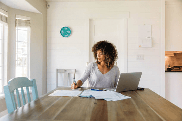 A woman sitting at a wooden table, focused on her finances while writing notes and working on a laptop in a bright, modern home environment.