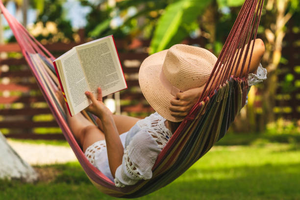 A woman relaxing in a hammock wearing a straw hat, reading a book, surrounded by greenery.