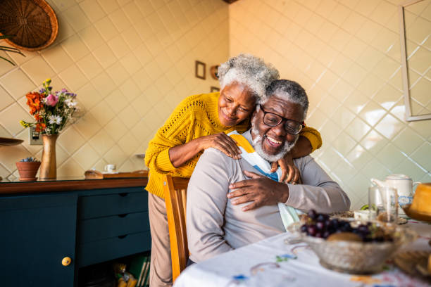 An elderly couple sharing a joyful moment at a dining table, with the woman affectionately embracing the man from behind, set in a cozy kitchen.