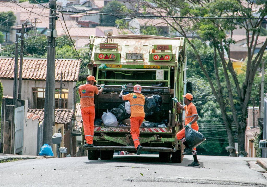 Three sanitation workers in orange uniforms collecting trash from a residential street, emptying garbage bags into a green garbage truck.