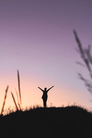 A silhouette of a person standing on a hilltop with arms outstretched, against a colorful sunset sky.