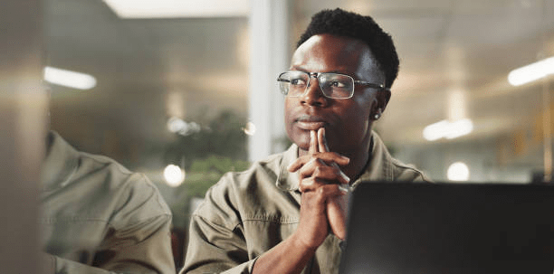 A young man sitting at a desk, looking thoughtfully with his hands clasped, reflecting on a decision, with a laptop in front of him.
