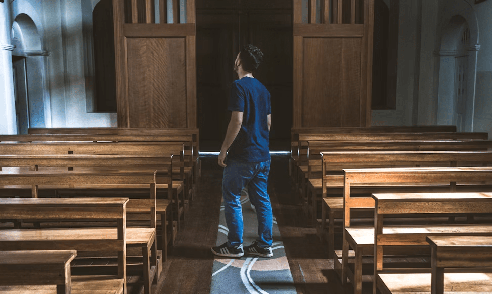 A person stands in an empty church, looking up towards the front, surrounded by wooden benches.