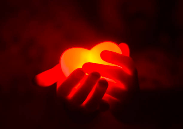 A pair of hands holding a glowing red heart in a dimly lit environment, symbolizing love and warmth.