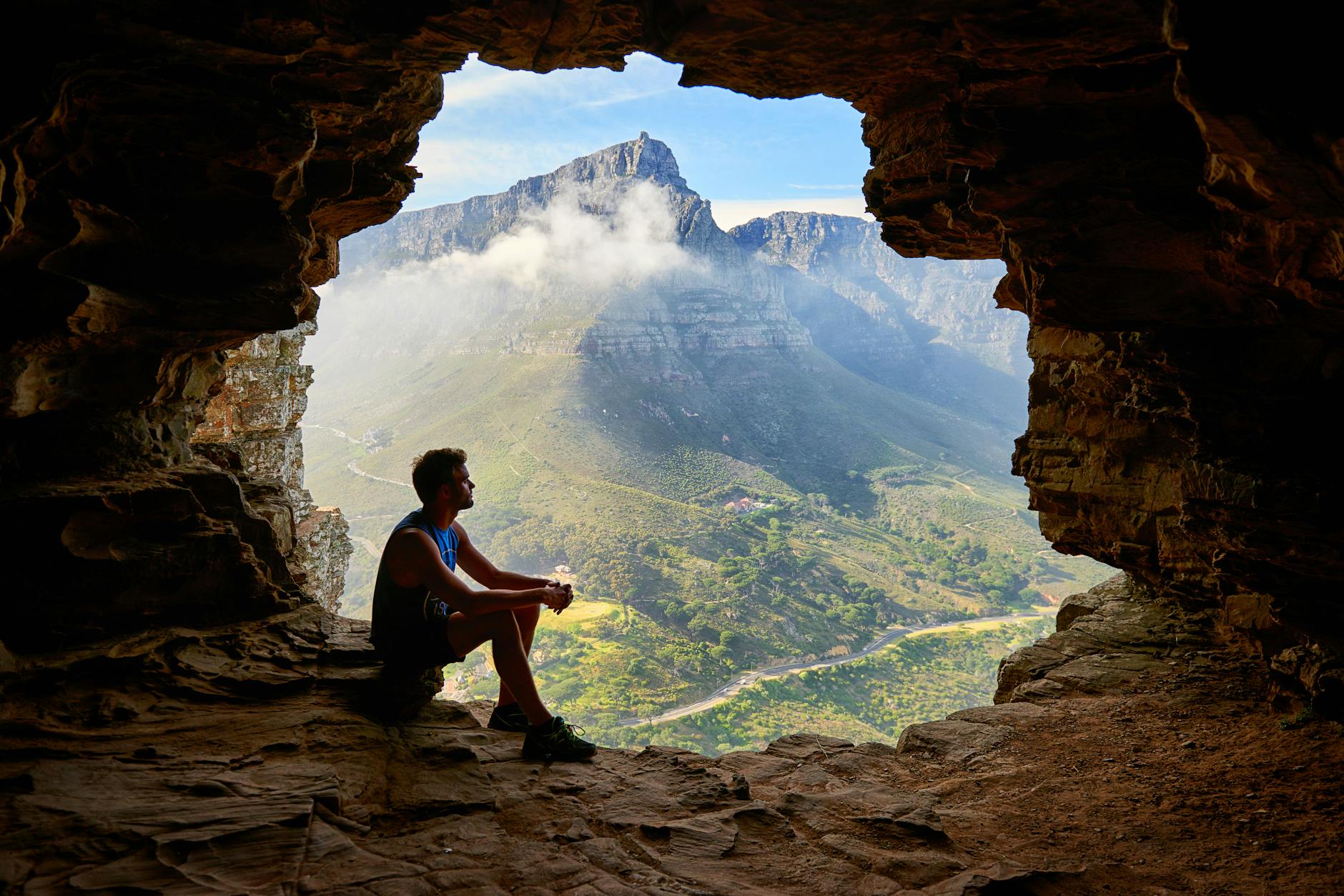 A person sitting inside a cave, looking out at a scenic mountain landscape with lush greenery and a winding road.