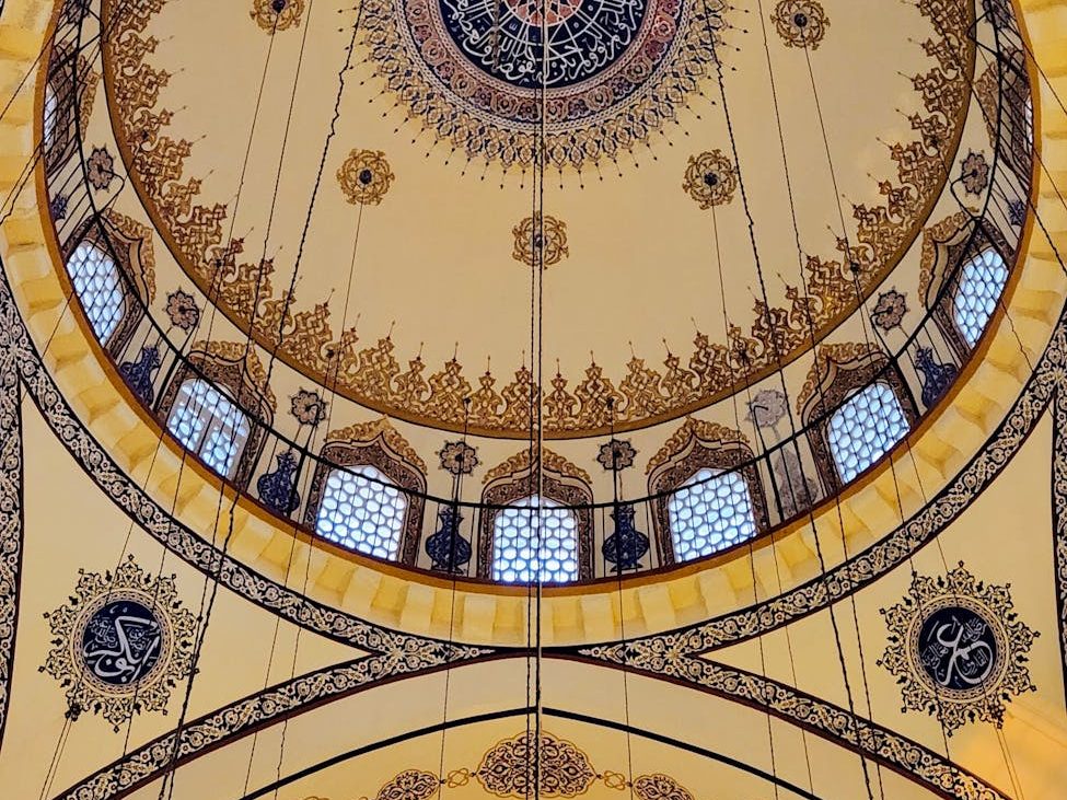 Interior view of a beautifully decorated mosque ceiling with intricate designs and patterns, featuring a circular dome and hanging lights.