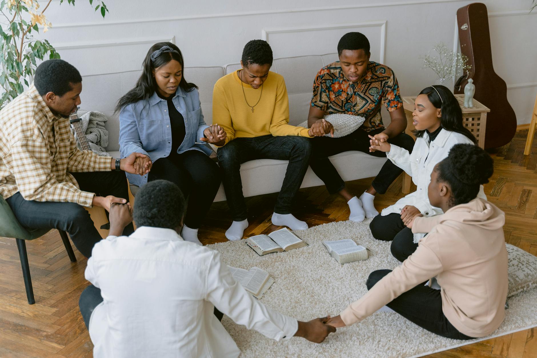 A group of six people sitting in a circle, holding hands and praying together on a carpet, with open Bibles in front of them.