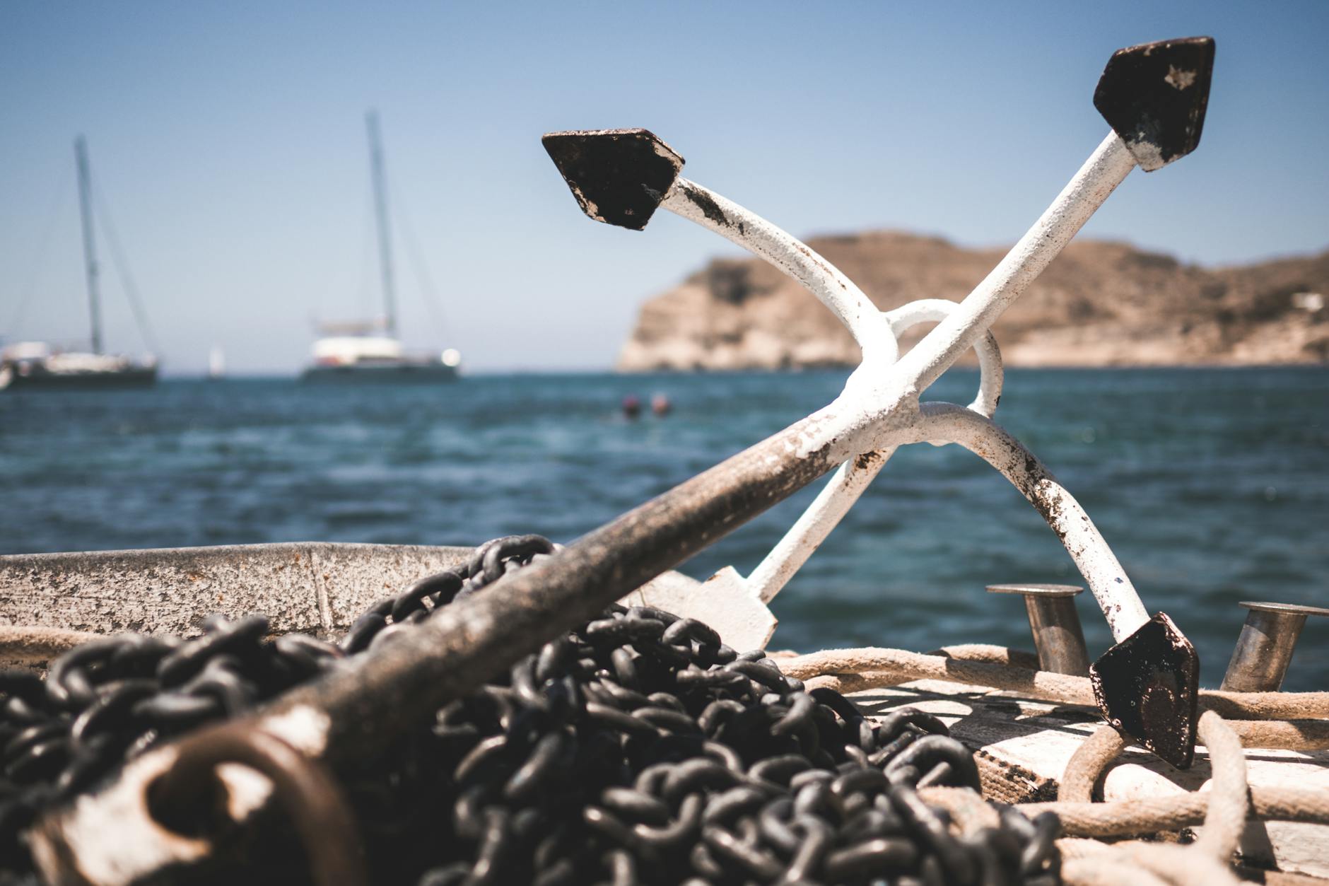 Close-up of a ship's anchor with a chain, set against a serene ocean and distant boats.