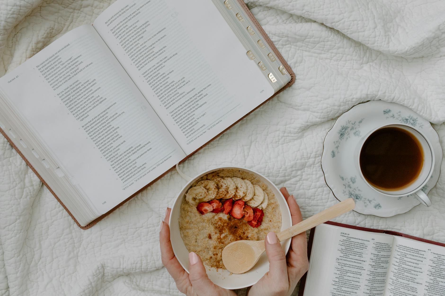 A person holding a bowl of oatmeal topped with banana and strawberries, with a wooden spoon, beside an open Bible and a cup of herbal drink on a white quilted surface.