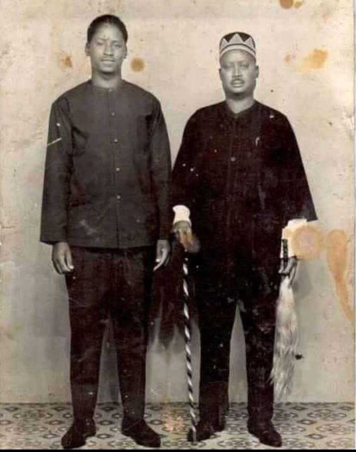 Historical black and white photograph of Raila and his father Oginga Odinga, side by side, with Oginga holding a ornate cane and wearing a traditional hat, indicative of cultural attire.
