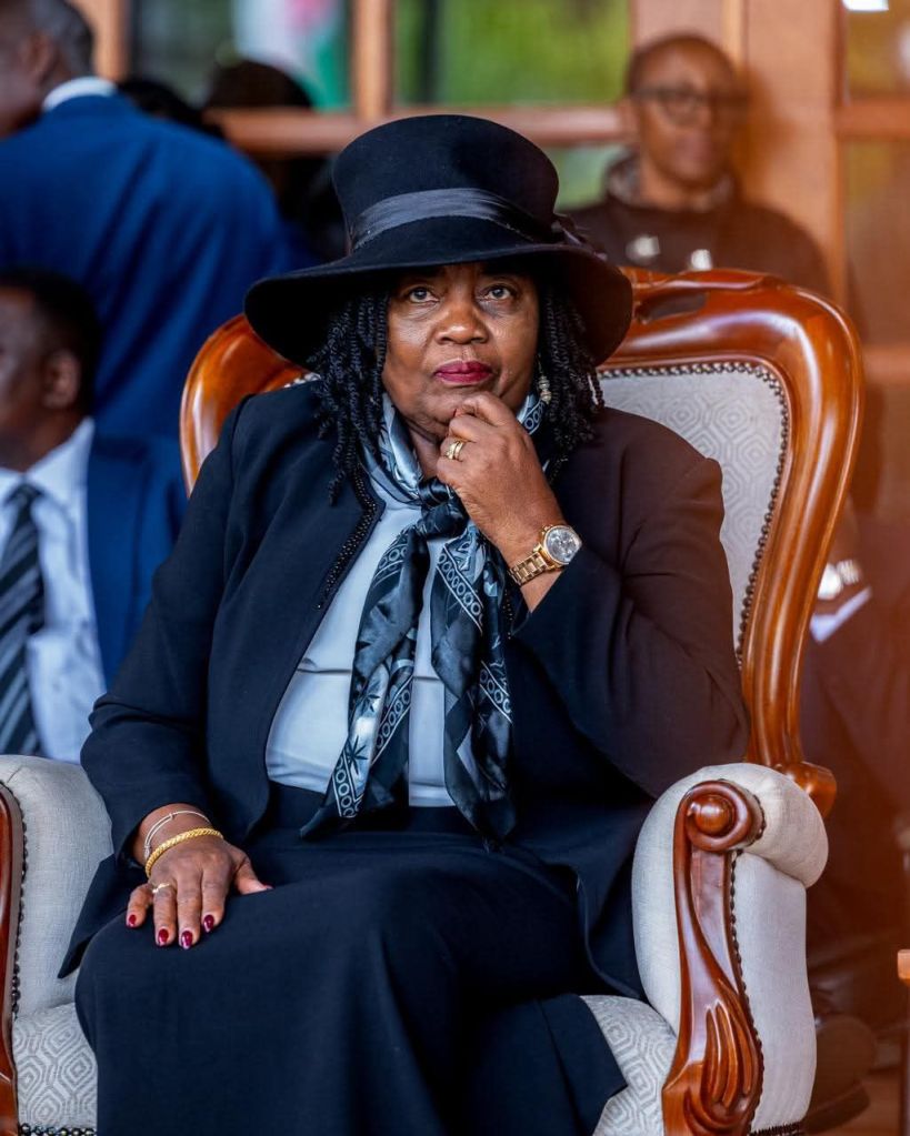 A woman in a black outfit and hat, seated in a wooden chair, with a thoughtful expression, during a solemn event.