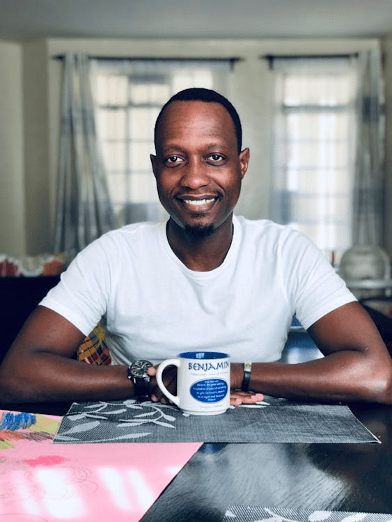 A smiling individual sitting at a table with a cup in hand, surrounded by a tidy indoor environment.