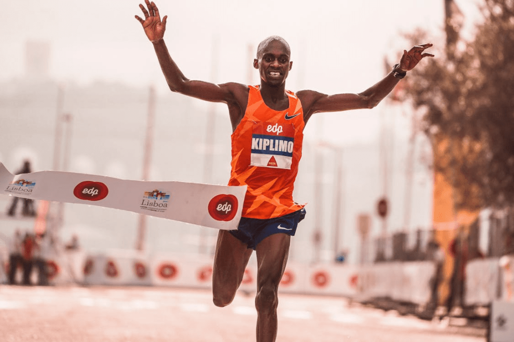 A male runner in an orange athletic outfit crosses the finish line with arms raised in celebration during a race.