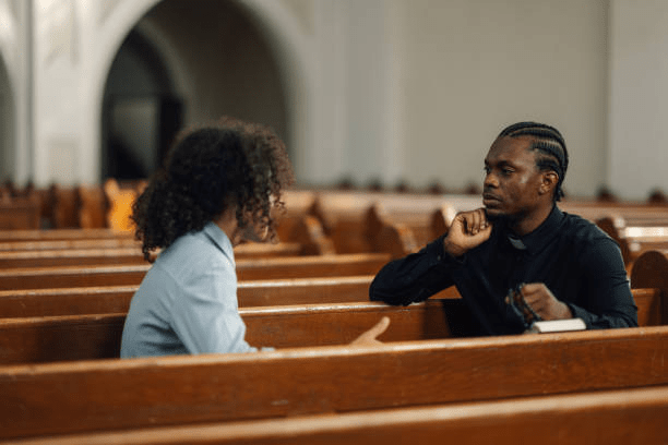 A man and a woman conversing inside a church, seated on wooden benches.